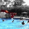 Piscina con scivolo rosso, molte persone nuotano e si divertono sotto il sole.