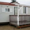 Una casa mobile bianca con tetto rosso e veranda in legno su terreno sabbioso.
