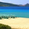 Spiaggia sabbiosa con mare cristallino, ombrelloni chiari e colline in lontananza.